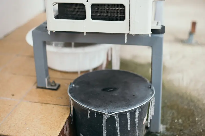Icicles hanging under a white outdoor unit raised on a metal stand, with ice running down into a black bucket placed below it